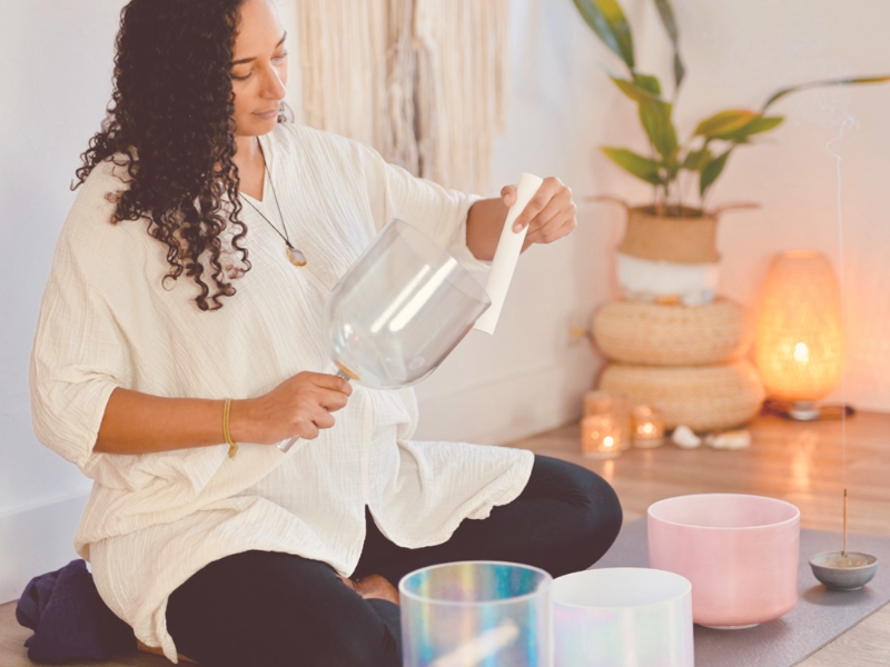 A woman in a white top and dark torusers sits on the floor holding a handhld crystal tones singing bowl and plays it with a white silicone sonic mallet. There are other crystal tones singing bowls infront of her.