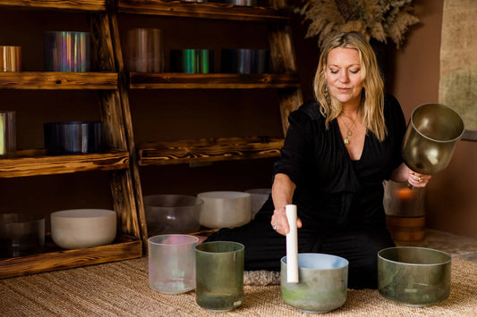 Woman - Kate - playing a set of singing bowls in a room with wooden shelves while looking down.
