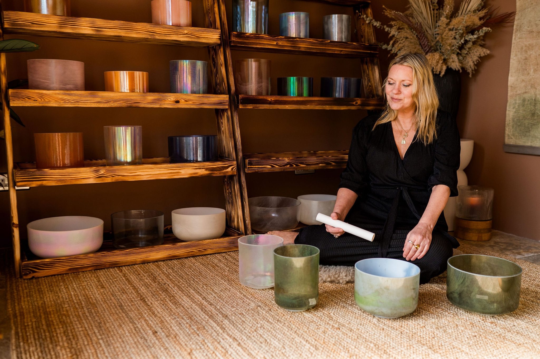 Woman sitting on a mat with singing bowls in a room with wooden shelves and decor.